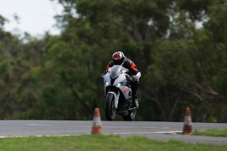 A motorcyclist wearing protective gear, including a helmet and a suit, is riding a white and black sport motorcycle on a track. The bike is slightly airborne, indicating high speed or a jump. The background consists of blurred green trees, and there are traffic cones near the track.
