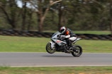 A motorcyclist wearing protective gear and a helmet is performing a wheelie on a sport motorcycle on a racetrack. The background consists of blurred greenery and tires, indicating a high-speed action shot.