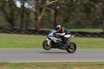 A motorcyclist wearing protective gear and a helmet is performing a wheelie on a sport motorcycle on a racetrack. The background consists of blurred greenery and tires, indicating a high-speed action shot.