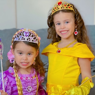 Two young children wearing princess costumes, including elaborate tiaras and jewelry. One is dressed in a yellow gown with golden gloves, while the other wears a purple dress with pink and purple accents. They are smiling and appear to be having fun indoors.