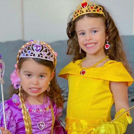 Two young children wearing princess costumes, including elaborate tiaras and jewelry. One is dressed in a yellow gown with golden gloves, while the other wears a purple dress with pink and purple accents. They are smiling and appear to be having fun indoors.