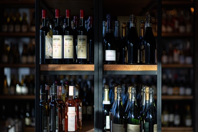 A variety of wine and champagne bottles are neatly arranged on dark wooden shelves. The scene is set in dim lighting, highlighting the bottles and emphasizing the labels. The background is slightly blurred, putting focus on the bottles themselves.