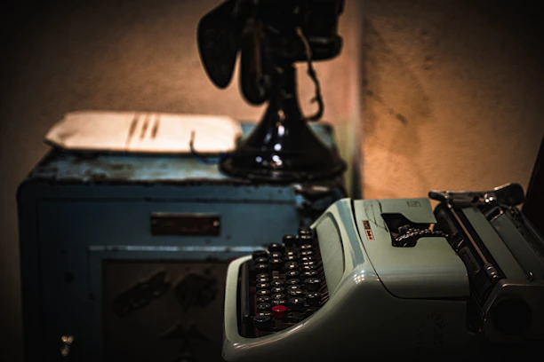 A dimly lit vintage typewriter on a dark wooden desk with scattered manuscript pages.