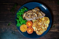 A blue plate features a visually appealing arrangement of food: grilled meat topped with sautéed mushrooms, crispy roasted potatoes with caramelized onions, fresh green kale, roasted garlic, and sliced tomatoes. The background is a rustic wooden surface with scattered greenery.