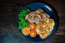 A blue plate features a visually appealing arrangement of food: grilled meat topped with saut&eacute;ed mushrooms, crispy roasted potatoes with caramelized onions, fresh green kale, roasted garlic, and sliced tomatoes. The background is a rustic wooden surface with scattered greenery.