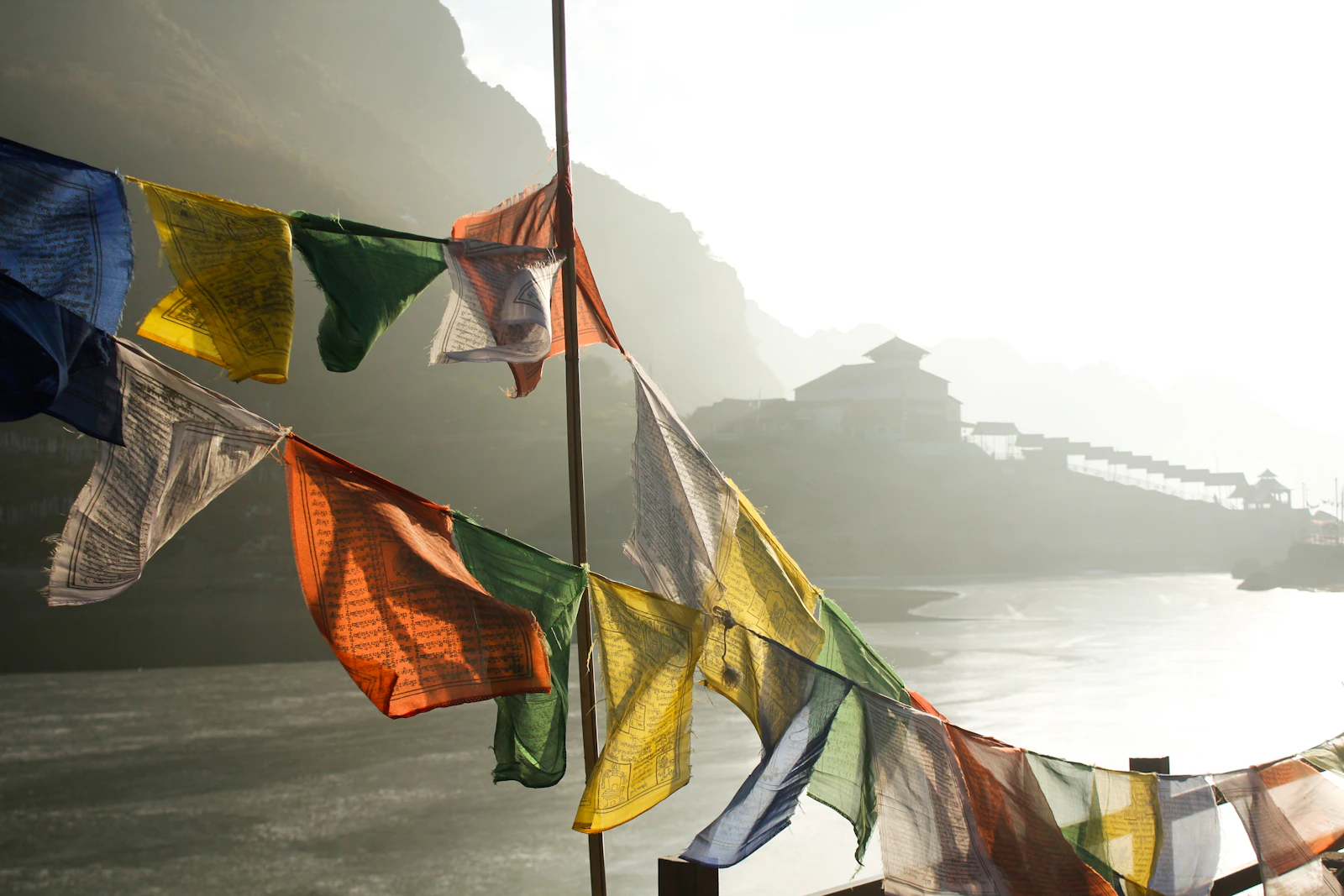 Prayer flags snapping in the wind beside Tsomgo Lake, hazy morning sun on the water and a lakeside building beyond