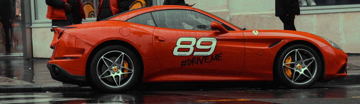 A vivid red sports car with the number 89 and the hashtag #DRIVEME painted on its side, parked on a wet urban street. The car's sleek design and alloy wheels stand out against the damp, rainy weather. People dressed in dark and red clothing are visible in the background.