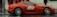A vivid red sports car with the number 89 and the hashtag #DRIVEME painted on its side, parked on a wet urban street. The car's sleek design and alloy wheels stand out against the damp, rainy weather. People dressed in dark and red clothing are visible in the background.