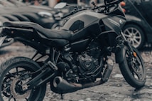 A sleek black motorcycle is parked on a wet surface, with visible raindrops on its body, emphasizing its shiny and pristine condition. The motorcycle features a robust engine and modern design, with a focus on its detailed mechanics. In the background, blurred images of cars add to the urban setting.