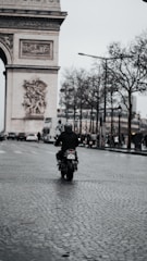 A lone motorcyclist rides down a cobblestone street in an urban setting with the Arc de Triomphe in the background. The sky is overcast, and bare trees line the street, with pedestrians and cars visible in the distance.