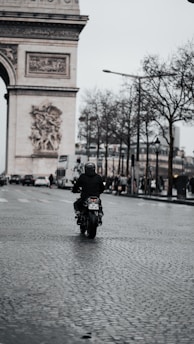 A lone motorcyclist rides down a cobblestone street in an urban setting with the Arc de Triomphe in the background. The sky is overcast, and bare trees line the street, with pedestrians and cars visible in the distance.