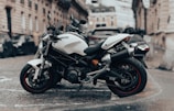 A sleek white motorcycle is parked on a cobblestone street in an urban environment. The motorcycle is positioned at an angle, showcasing its modern design and sporty features. In the background, there is a blurred view of historic European-style buildings and other parked vehicles, suggesting a city setting.