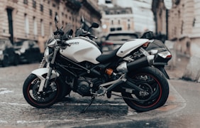A sleek white motorcycle is parked on a cobblestone street in an urban environment. The motorcycle is positioned at an angle, showcasing its modern design and sporty features. In the background, there is a blurred view of historic European-style buildings and other parked vehicles, suggesting a city setting.