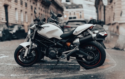 A sleek white motorcycle is parked on a cobblestone street in an urban environment. The motorcycle is positioned at an angle, showcasing its modern design and sporty features. In the background, there is a blurred view of historic European-style buildings and other parked vehicles, suggesting a city setting.