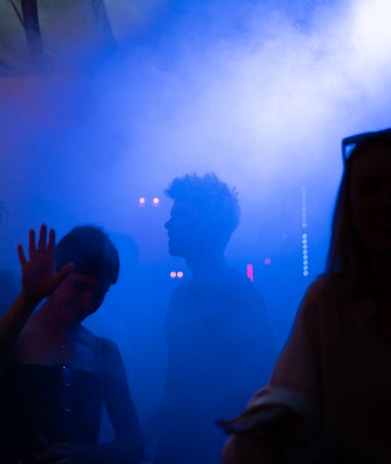 Silhouettes of people are visible in a dimly lit environment filled with blue fog or smoke. The atmosphere appears to be lively, possibly at a nightclub or concert, with faint lights in the background.