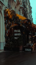 A street view of a cafe with signs offering pizza, burgers, and pasta. The cafe has a yellow awning adorned with lush greenery and flowers. The building features ornate architecture with detailed stonework. Outdoor seating is visible with stacked chairs. The overall scene appears moody due to the overcast weather.