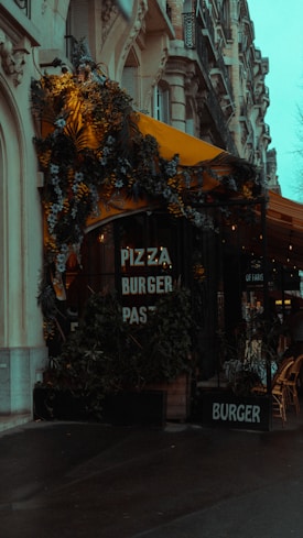 A street view of a cafe with signs offering pizza, burgers, and pasta. The cafe has a yellow awning adorned with lush greenery and flowers. The building features ornate architecture with detailed stonework. Outdoor seating is visible with stacked chairs. The overall scene appears moody due to the overcast weather.