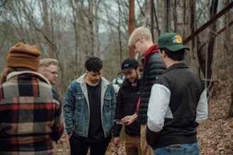 A group of people is gathered in a wooded area, dressed in casual, warm clothing. They appear to be focused on a smartphone that one person is holding. The background is a forest with bare trees, suggesting a chilly or early spring day.