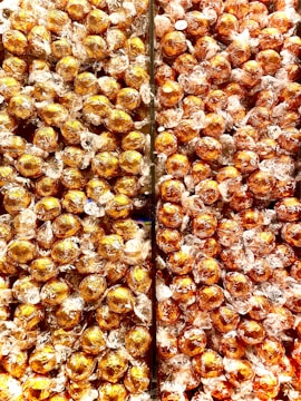 A large quantity of round chocolates wrapped in shiny golden foil, tightly packed together in a display. Each chocolate is individually wrapped with transparent materials around the gold foil.