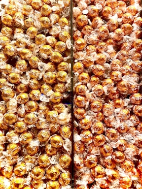 A large quantity of round chocolates wrapped in shiny golden foil, tightly packed together in a display. Each chocolate is individually wrapped with transparent materials around the gold foil.