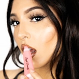 A close-up of a person with carefully styled dark hair and dramatic makeup, including dark eyeliner and glossy lips. A pink tube of lip gloss with the brand name 'Loyal Beauty' is held near the lips.