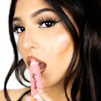 A close-up of a person with carefully styled dark hair and dramatic makeup, including dark eyeliner and glossy lips. A pink tube of lip gloss with the brand name 'Loyal Beauty' is held near the lips.