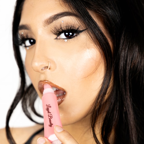 A close-up of a person with carefully styled dark hair and dramatic makeup, including dark eyeliner and glossy lips. A pink tube of lip gloss with the brand name 'Loyal Beauty' is held near the lips.