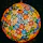 A spherical lampshade composed of intricate floral patterns in vibrant colors, with a mosaic of red, orange, blue, green, and yellow elements. The design allows light to diffuse through the gaps, creating a warm and colorful glow.