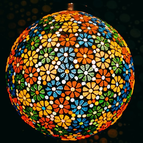 A spherical lampshade composed of intricate floral patterns in vibrant colors, with a mosaic of red, orange, blue, green, and yellow elements. The design allows light to diffuse through the gaps, creating a warm and colorful glow.