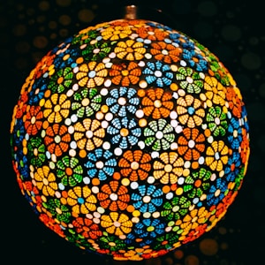 A spherical lampshade composed of intricate floral patterns in vibrant colors, with a mosaic of red, orange, blue, green, and yellow elements. The design allows light to diffuse through the gaps, creating a warm and colorful glow.