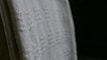 A close-up view of a white textured fabric with intricate woven patterns, displayed against a dark background. The fabric edges appear detailed and the weave texture is clearly visible, emphasizing its tactile quality.