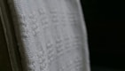 A close-up view of a white textured fabric with intricate woven patterns, displayed against a dark background. The fabric edges appear detailed and the weave texture is clearly visible, emphasizing its tactile quality.
