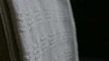 A close-up view of a white textured fabric with intricate woven patterns, displayed against a dark background. The fabric edges appear detailed and the weave texture is clearly visible, emphasizing its tactile quality.
