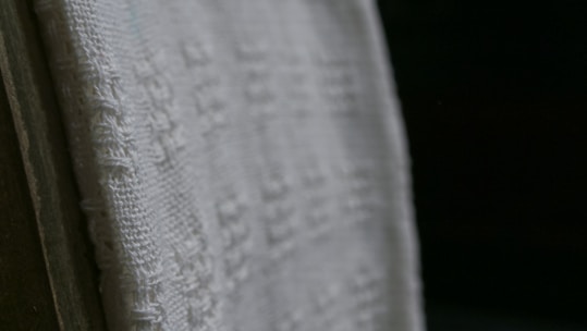 A close-up view of a white textured fabric with intricate woven patterns, displayed against a dark background. The fabric edges appear detailed and the weave texture is clearly visible, emphasizing its tactile quality.