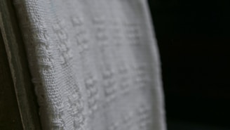 A close-up view of a white textured fabric with intricate woven patterns, displayed against a dark background. The fabric edges appear detailed and the weave texture is clearly visible, emphasizing its tactile quality.