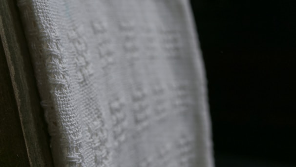 A close-up view of a white textured fabric with intricate woven patterns, displayed against a dark background. The fabric edges appear detailed and the weave texture is clearly visible, emphasizing its tactile quality.