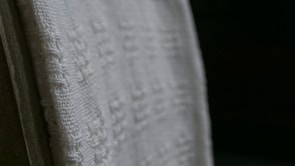 A close-up view of a white textured fabric with intricate woven patterns, displayed against a dark background. The fabric edges appear detailed and the weave texture is clearly visible, emphasizing its tactile quality.
