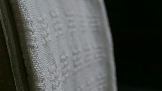 A close-up view of a white textured fabric with intricate woven patterns, displayed against a dark background. The fabric edges appear detailed and the weave texture is clearly visible, emphasizing its tactile quality.
