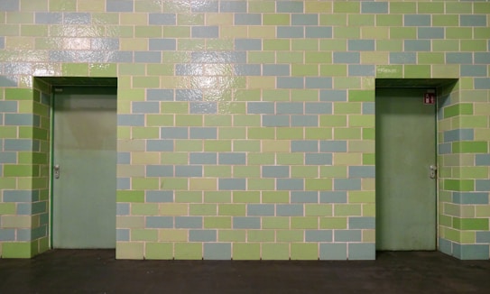 A wall made of rectangular tiles in alternating light green and pale blue colors, featuring two closed doors painted in a similar green tone. The tiles are arranged in a horizontal pattern, evenly distributing the colors across the surface.