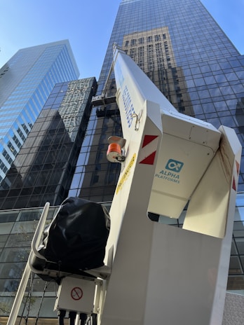Aerial lift equipment is positioned near a glass skyscraper, with reflections of neighboring buildings visible on the facade. The lift is marked with 'Alpha Platforms' and has safety warnings displayed.