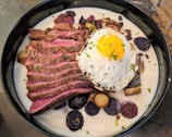 A dish featuring sliced medium-rare steak served in a creamy sauce with a fried egg on top. Surrounding the steak and egg are slices of colorful potatoes, including purple varieties, garnished with chives.