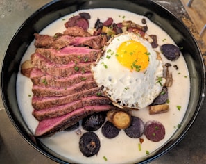 A dish featuring sliced medium-rare steak served in a creamy sauce with a fried egg on top. Surrounding the steak and egg are slices of colorful potatoes, including purple varieties, garnished with chives.