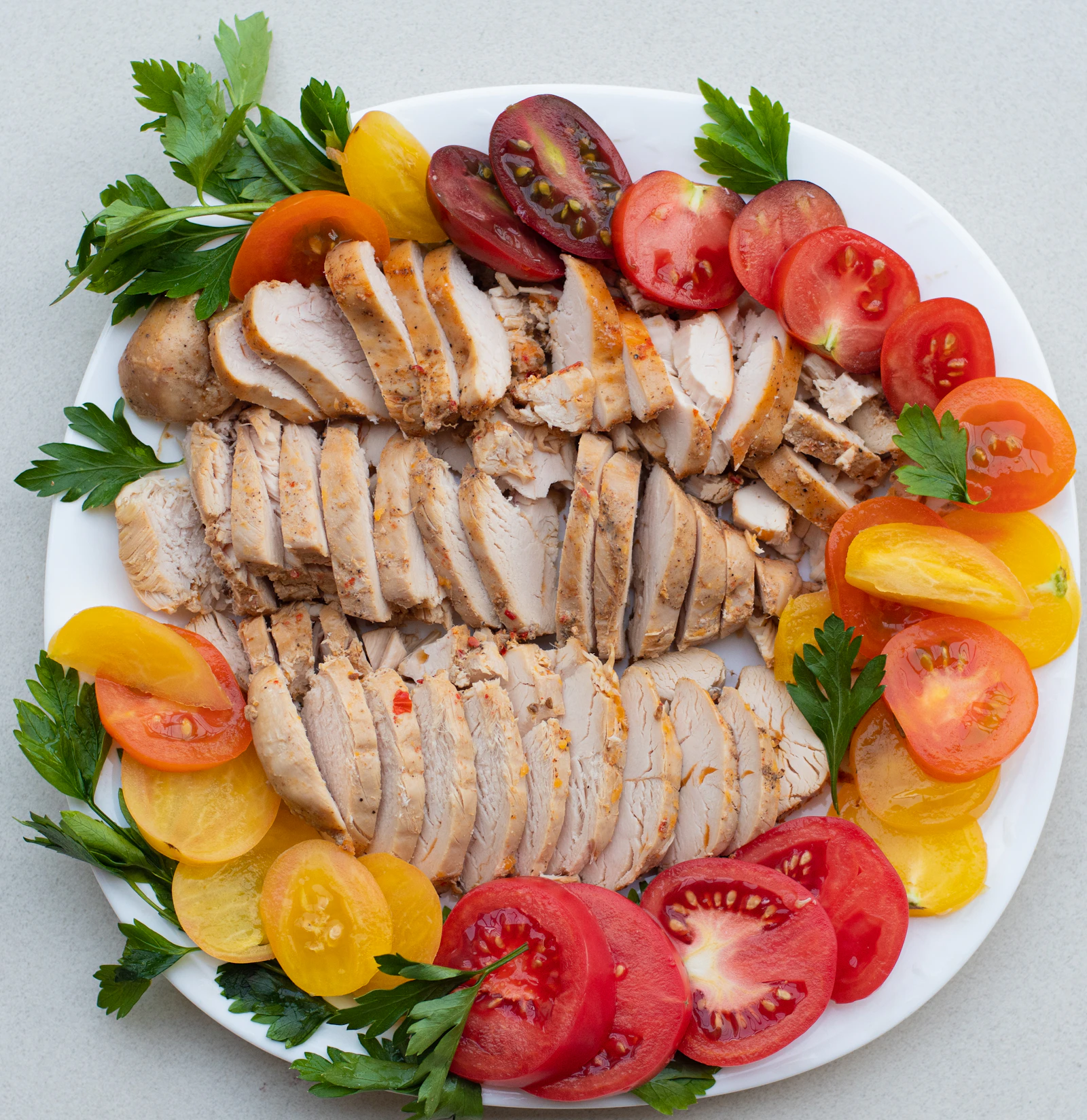 Sliced cooked chicken breast fanned across a plate and ringed with tomato wedges and parsley