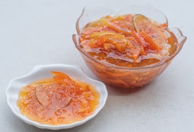 A close-up view of a small glass bowl and a ceramic dish, both filled with glossy orange marmalade, featuring pieces of fruit peel. The marmalade appears homemade, with vibrant orange slices visible and a thick, syrupy texture.