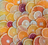 A vibrant assortment of sliced citrus fruits, including oranges, lemons, limes, and grapefruits, arranged in an overlapping pattern. The slices display a variety of colors from deep red to bright yellow and green, showcasing the diverse textures and juices of the fruits.
