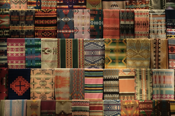 A collection of colorful woven fabrics featuring a variety of traditional geometric patterns. The fabrics are neatly arranged in rows, showcasing an array of intricate designs, including diamonds, zigzags, and stripes in diverse colors.