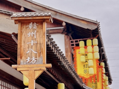 A wooden sign with Chinese characters stands prominently in front of a traditional-style building with a tiled roof. Yellow lanterns hang around the building, adding vibrant pops of color.