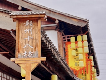 A wooden sign with Chinese characters stands prominently in front of a traditional-style building with a tiled roof. Yellow lanterns hang around the building, adding vibrant pops of color.