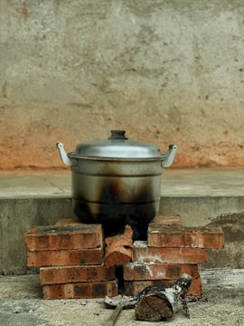 A metal pot with handles is placed on a makeshift stove made of stacked bricks. Below the pot, there are pieces of wood with signs of burning. The background features a concrete wall with a rough texture.