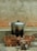 A metal pot with handles is placed on a makeshift stove made of stacked bricks. Below the pot, there are pieces of wood with signs of burning. The background features a concrete wall with a rough texture.